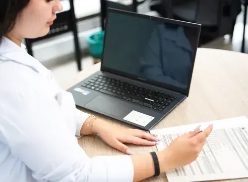 Mujer firmando unos documentos con un portátil a su lado.