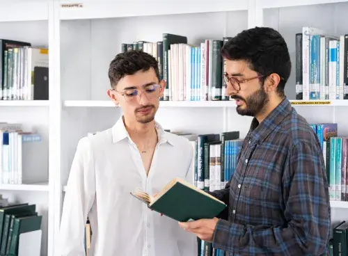 Dos personas observan un libro juntas mientras, en el fondo, se ve un estante lleno de libros.