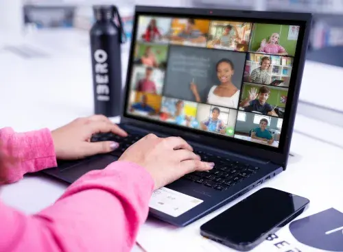 Persona frente a un portátil participando en una reunión virtual; al lado del equipo hay un celular y un termo.