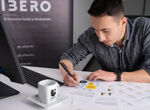 Persona con un bolígrafo en la mano revisando documentos sobre la mesa; a un lado hay un portátil.